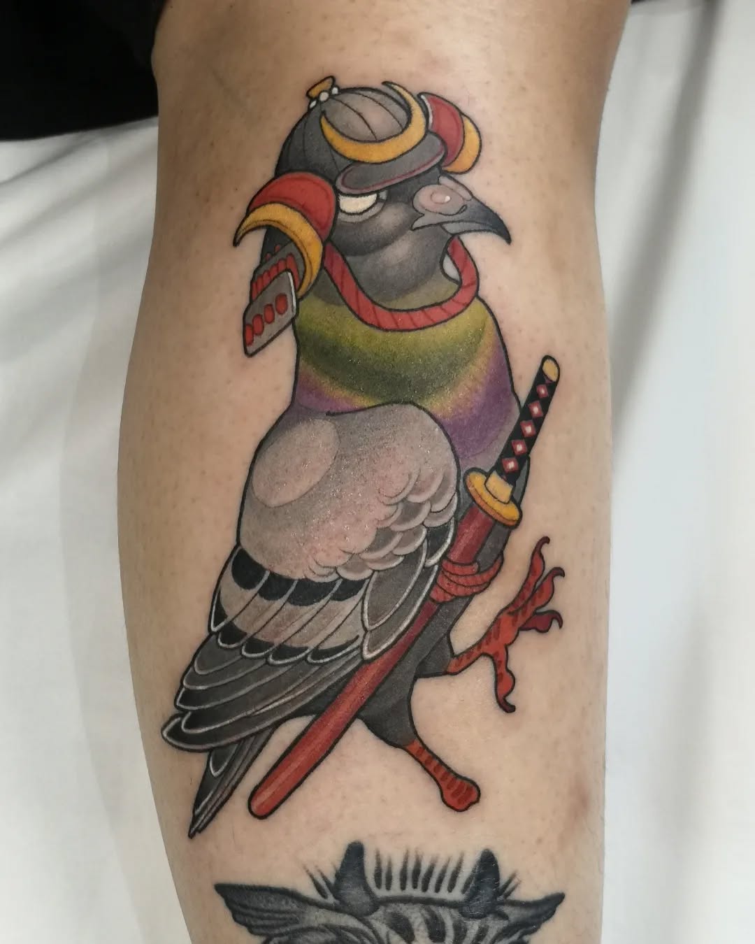 Colorful bird warrior character wearing armor and holding a red sword, tattooed on the leg with gold, red, purple, and grey tones.