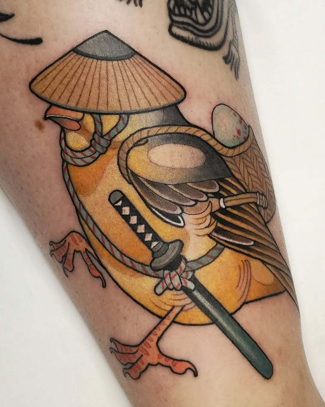Colorful tattoo on an arm featuring a bird wearing a conical hat and holding a sword, rendered in yellow, orange, and brown tones.
