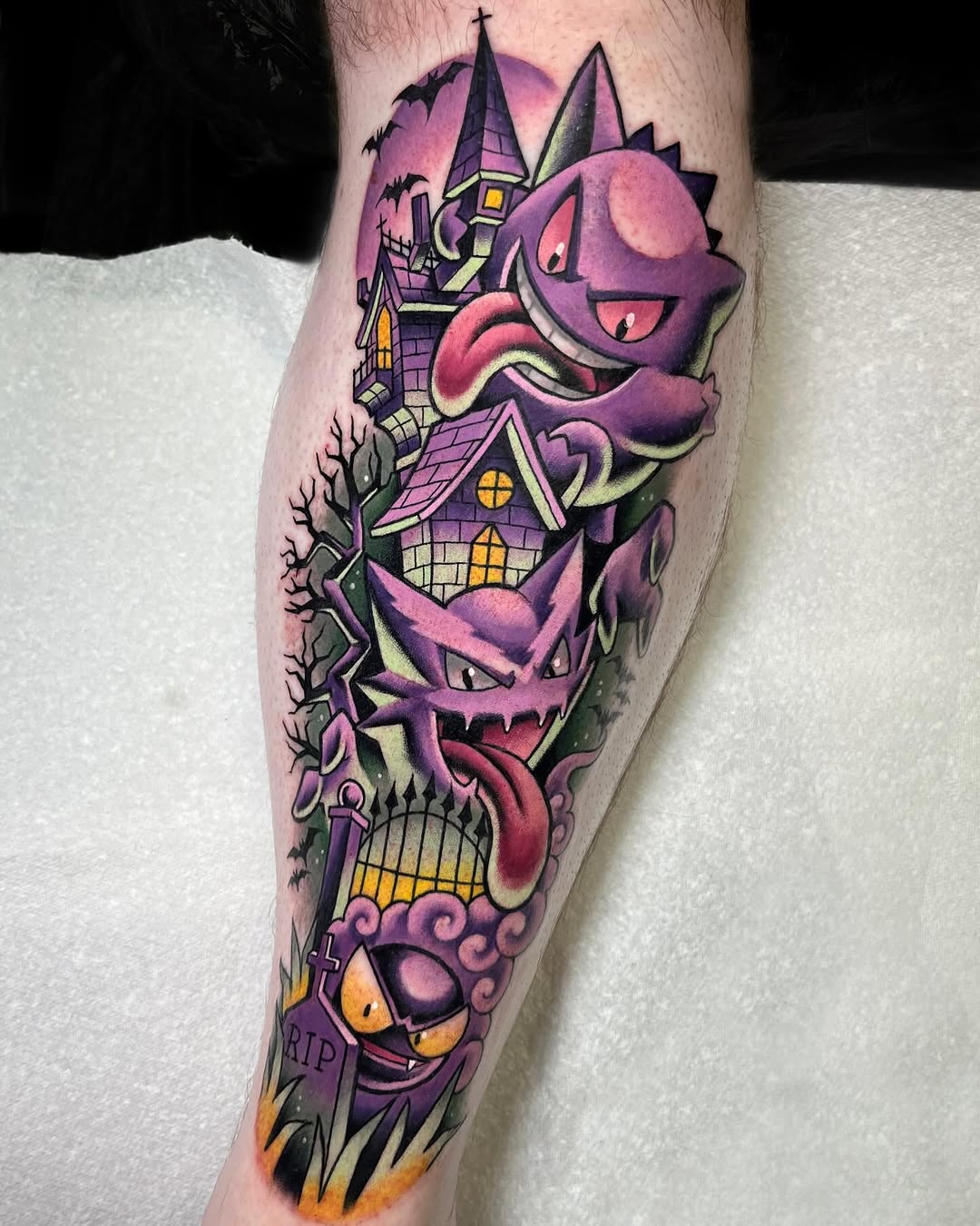 Colorful leg tattoo featuring stacked purple Pokémon characters with a haunted mansion, bare tree, gravestones, and pink sky background.