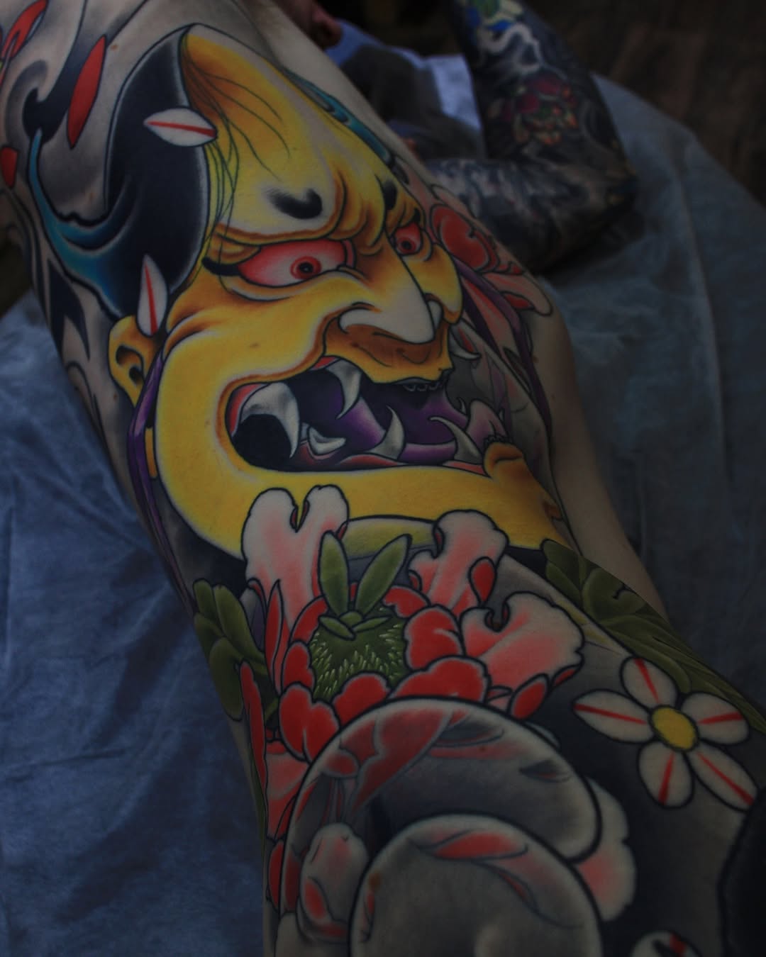 Colorful arm tattoo featuring a yellow demon face with red eyes and open mouth, surrounded by red and white flowers with green leaves.
