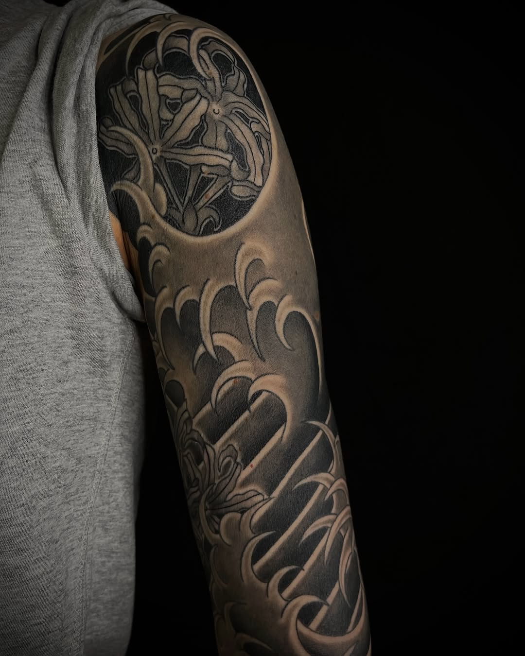 Full sleeve tattoo on upper arm featuring ornate circular design at shoulder with flowing scrollwork patterns extending down to elbow in black and grey ink.