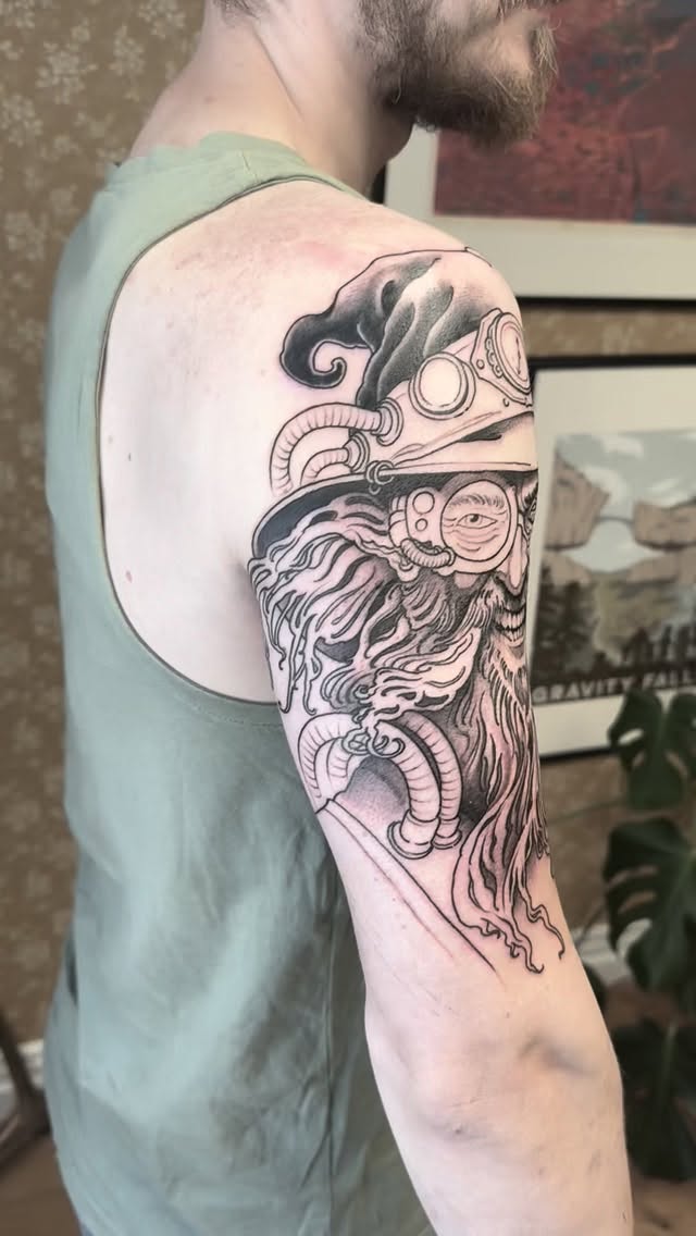 Detailed monochrome shoulder tattoo featuring a bearded face with ornamental scrollwork and decorative elements in black ink on pale skin.