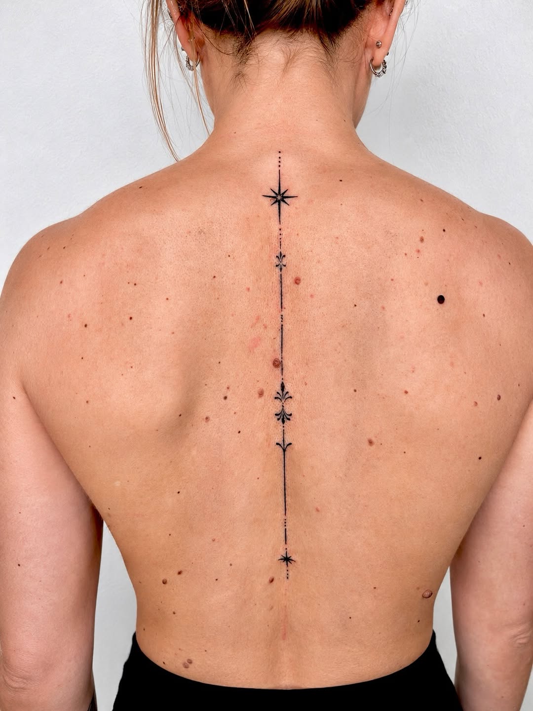 Vertical spine tattoo featuring black stars and ornamental flourishes running from the upper back to the lower back.
