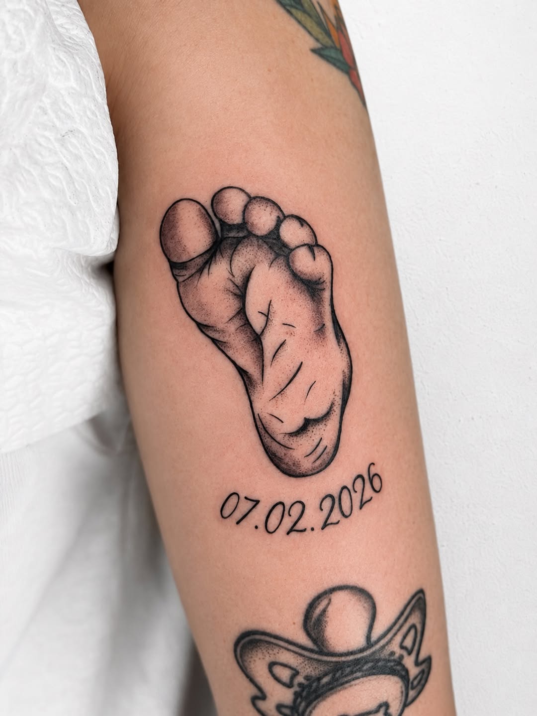 Baby footprint tattoo with date 07.02.2026 in black ink on the inner arm, positioned between other tattoos.