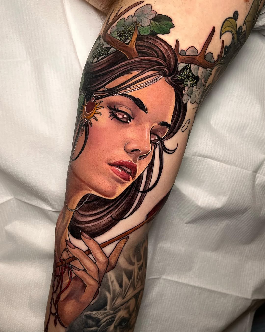 Female portrait tattoo on leg featuring brown hair, antlers, flowers, and hand gesture in warm tones of orange, brown, and green.