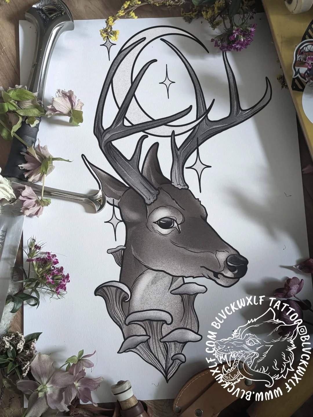 Detailed pen sketch of a deer head with large antlers, decorative mushroom elements below, and four-pointed stars, displayed on white paper surrounded by fresh flowers.