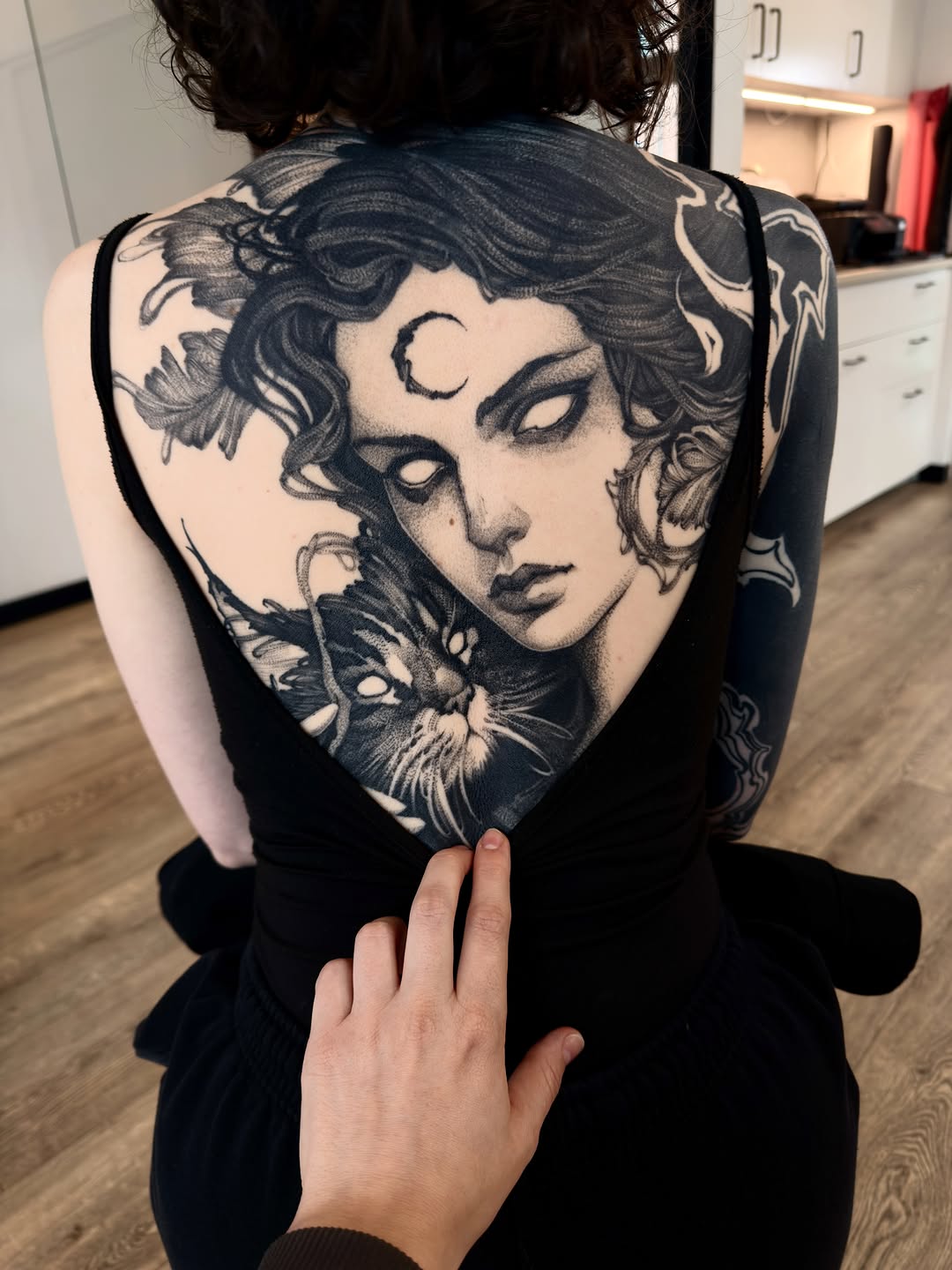 Large-scale portrait tattoo on upper back featuring a woman's face with closed eyes, crescent moon on forehead, and cat face integrated below, rendered in black ink.