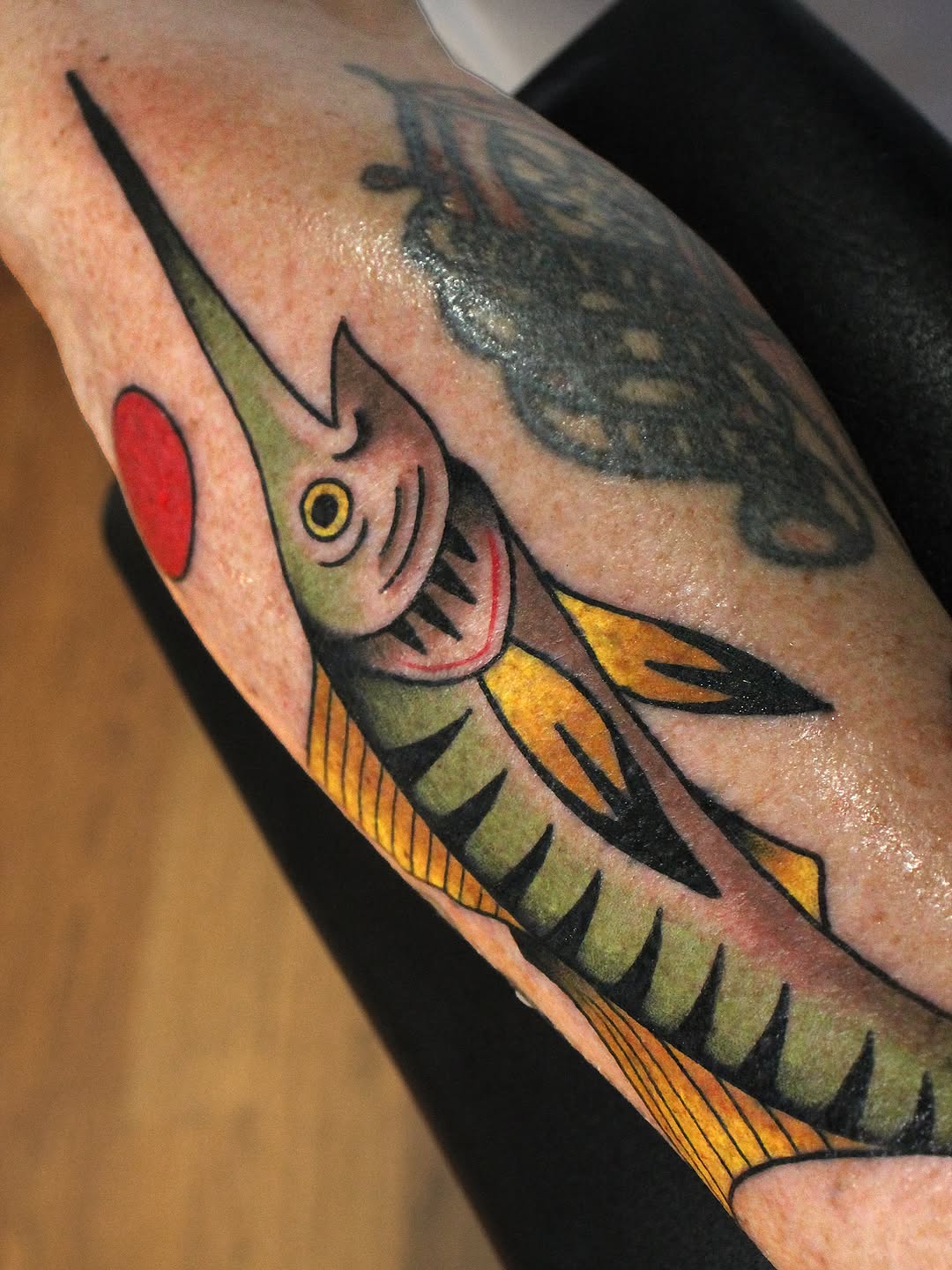 Colorful fish head tattoo on forearm featuring green, yellow, orange, and red tones with yellow eye and detailed gill markings.