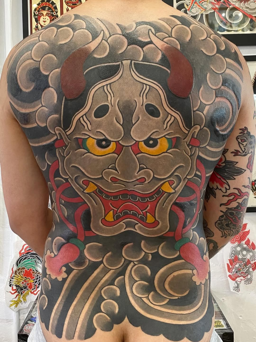 Large back tattoo featuring a beige and red demon mask with gold eyes and ornamental cloud patterns in gray and tan tones.