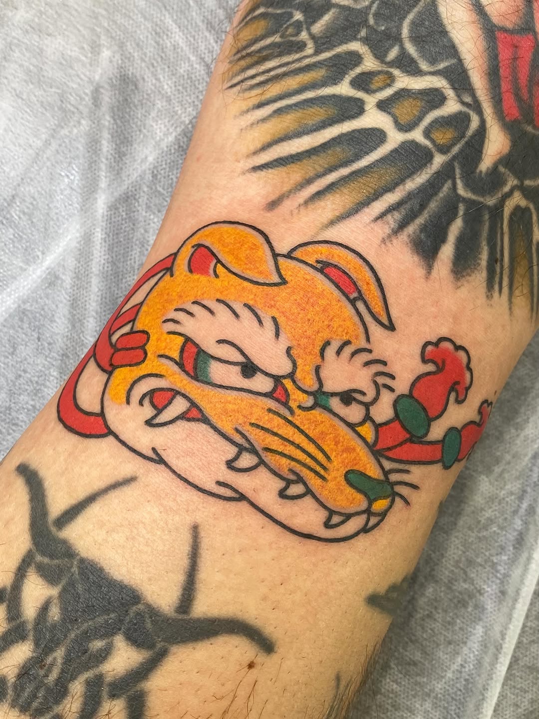 Golden cat figurine with red accents and raised paw tattoo on forearm, featuring black outlines and green details.
