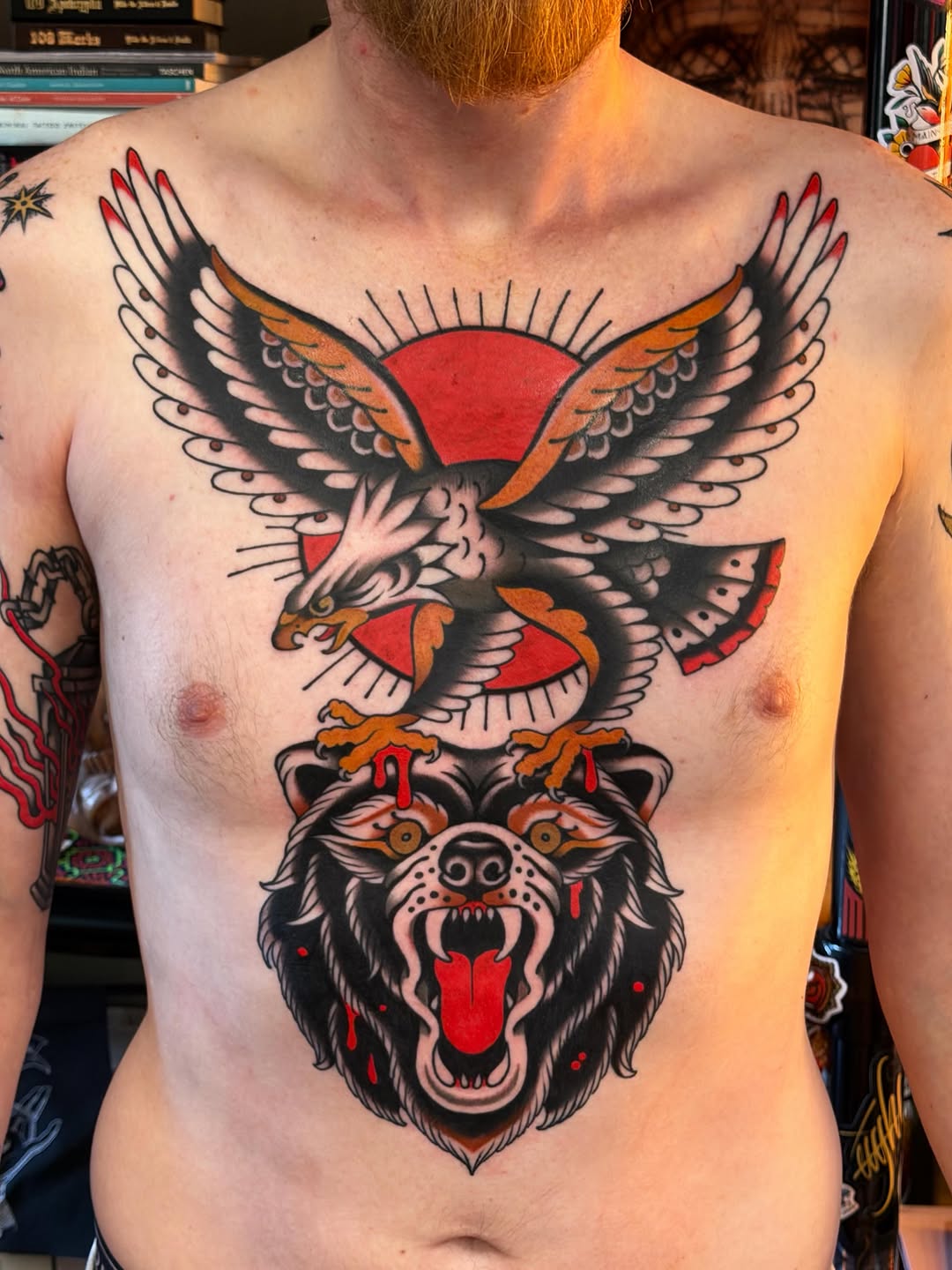 Chest tattoo featuring a red-winged eagle perched above a snarling bear head, rendered in black outlines with red and orange accents.