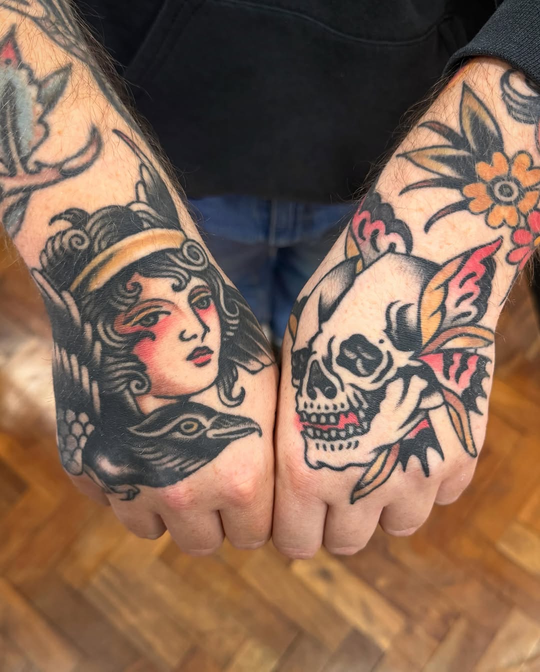 Two hands with matching tattoos featuring a winged female face on the left and a skull with flowers on the right, rendered in black, red, and yellow ink.