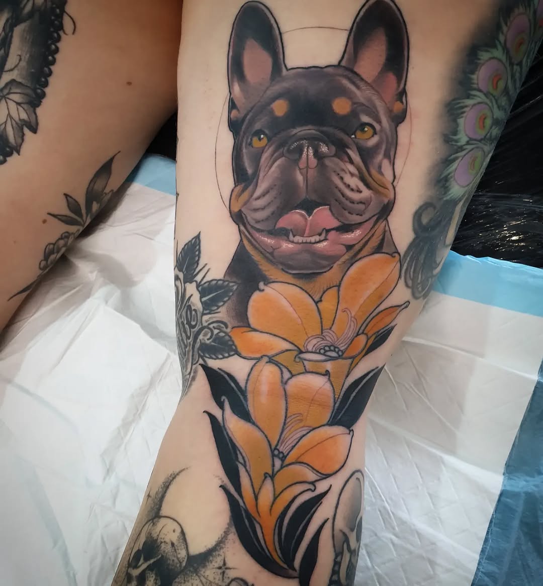 Colorful arm tattoo featuring a smiling black and tan French Bulldog head with golden eyes, surrounded by orange flowers down the forearm.