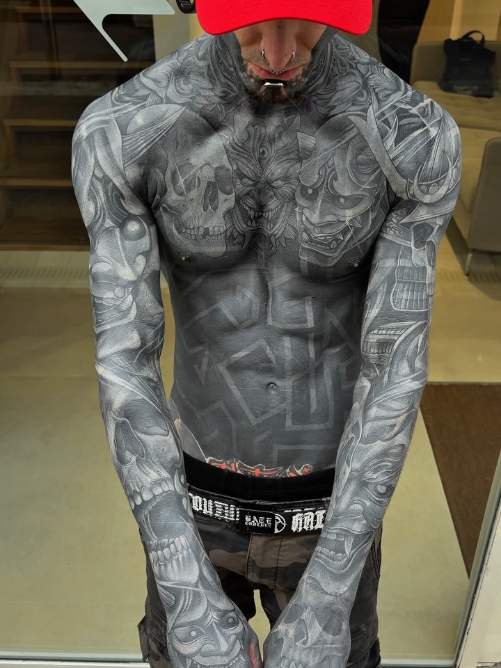 Man displays extensive gray-toned tattoos covering chest, shoulders, and both arms with skulls and ornamental designs in a portfolio photo.