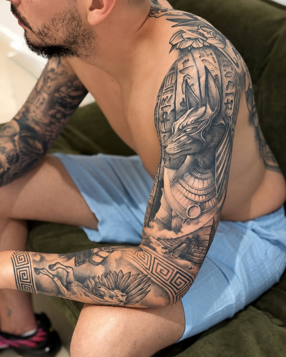 Shirtless man displaying full arm tattoos featuring an Egyptian deity head, hieroglyphics, floral elements, and geometric patterns in black ink.