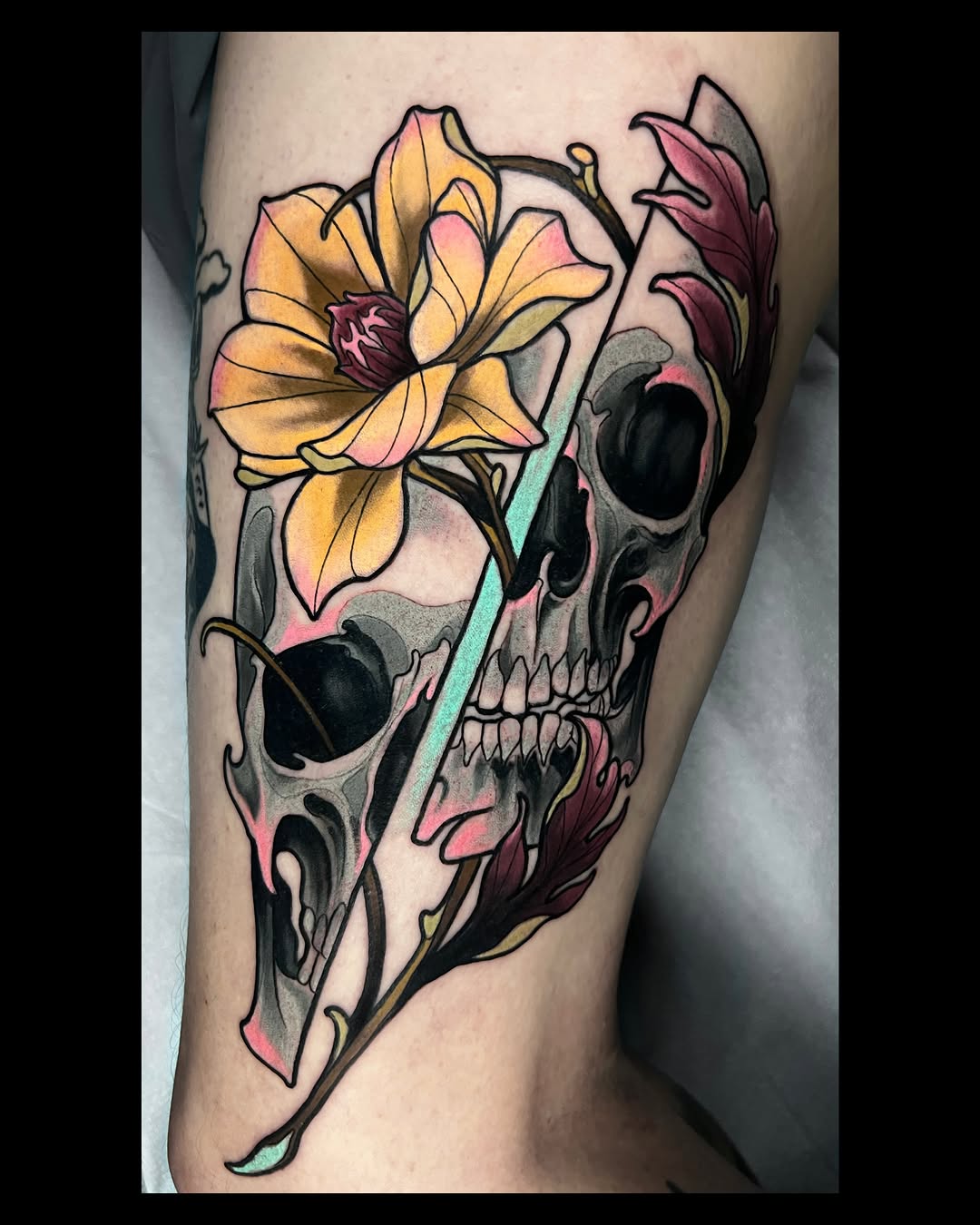 Colorful skull tattoo with yellow and pink flower on upper leg, featuring black outlines and gradient shading in multiple colors.