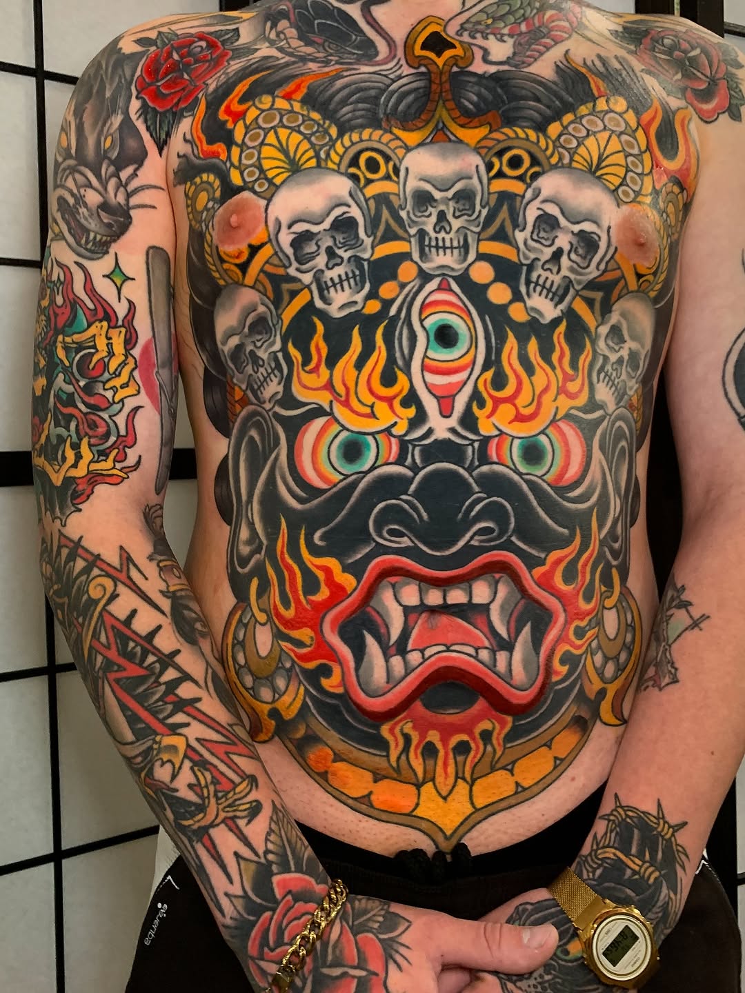 Colorful chest and sleeve tattoo featuring skulls, flames, ornamental designs, roses, and mystical eyes in red, orange, gold, and black.