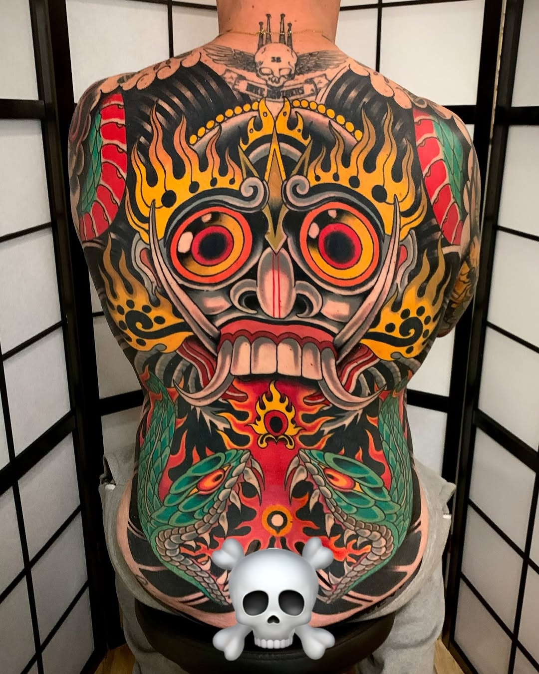 Colorful back tattoo featuring a large skull with red eyes and flame crown, winged insects below, and decorative elements in red, yellow, green, and black.
