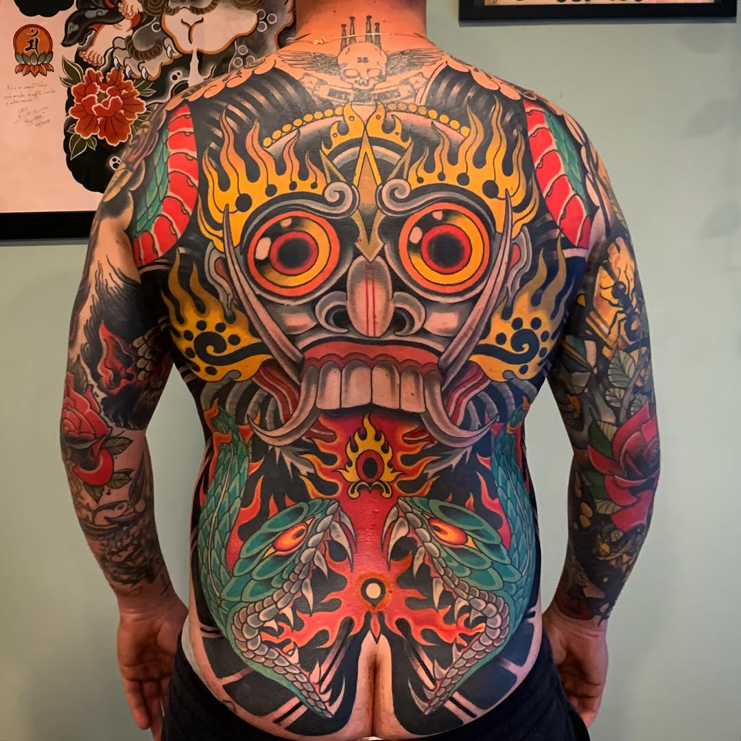 Back tattoo featuring a large ornate owl mask with red and gold eyes, surrounded by flames, flowers, and decorative motifs in vibrant red, orange, yellow, teal, and black.