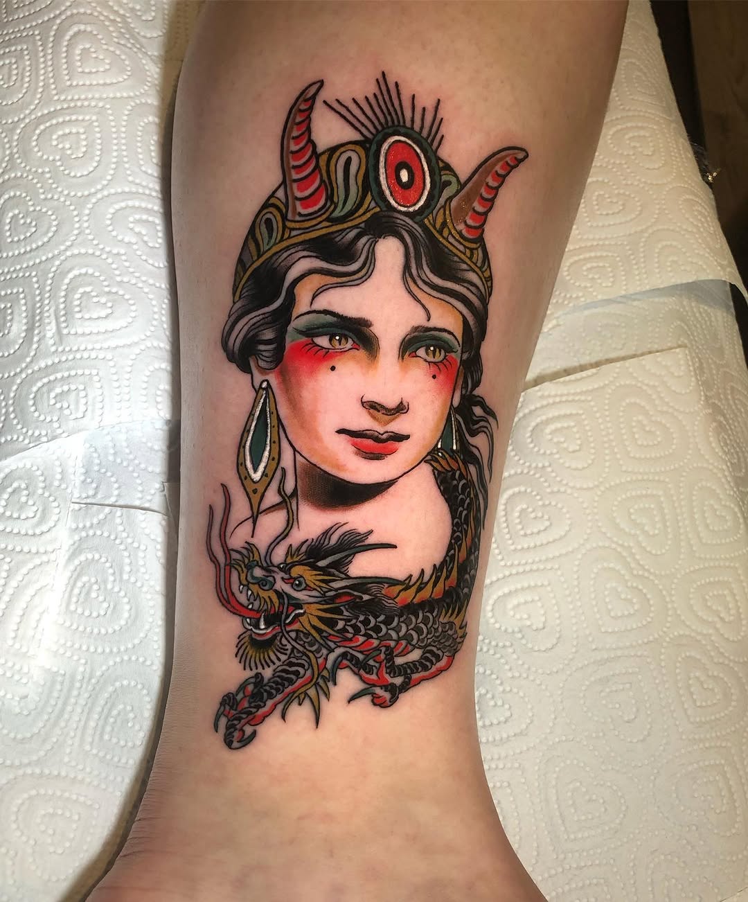 Colorful portrait tattoo on forearm featuring a woman with horns, red eyes, braided hair, dragon wrapped around neck, and decorative earrings against skin.