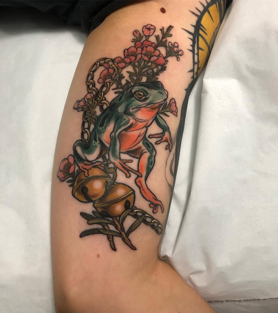 Colorful frog tattoo with red flowers and botanical elements on upper arm, featuring green, orange, and red ink.