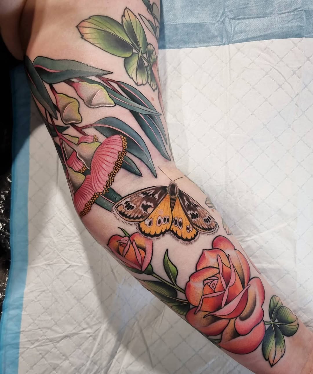 Colorful arm tattoo featuring roses, a butterfly, and botanical elements in red, orange, yellow, and green on the forearm and upper arm.
