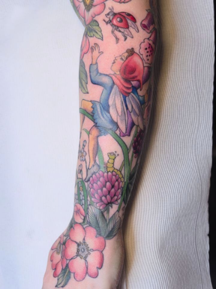 Colorful full-sleeve tattoo featuring flowers, ladybugs, a fairy figure, and botanical elements in pink, blue, and green on the forearm and arm.