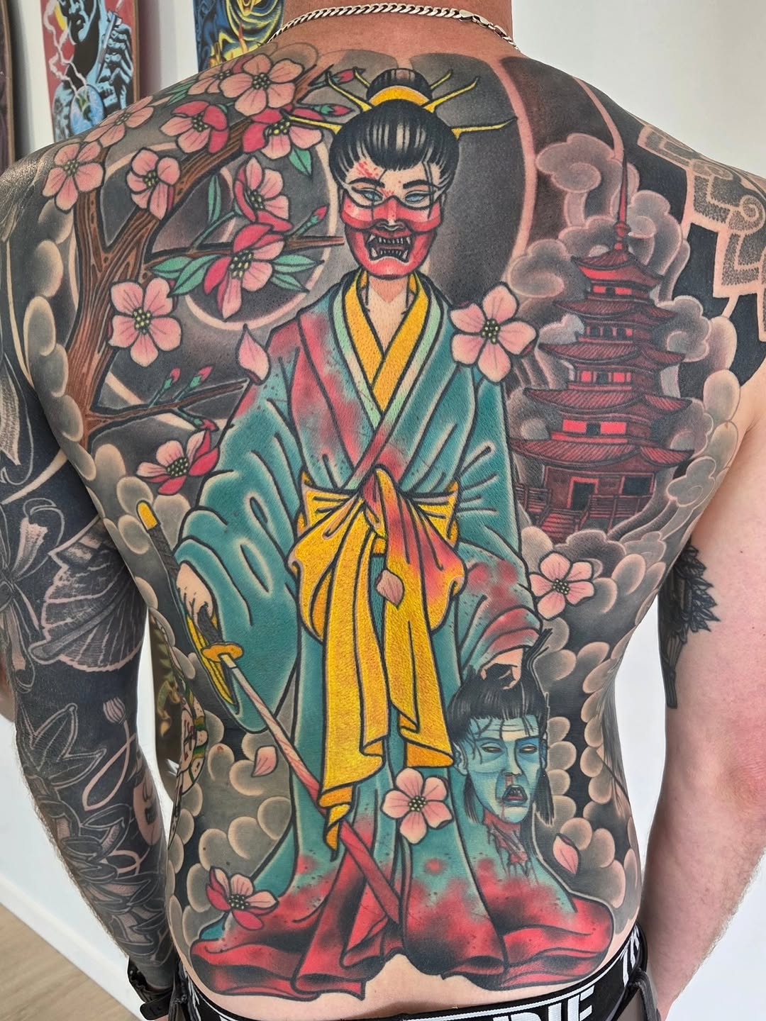 Large back tattoo featuring a demon-faced figure in teal and yellow robes surrounded by pink flowers, a pagoda, clouds, and a smaller demon head below.