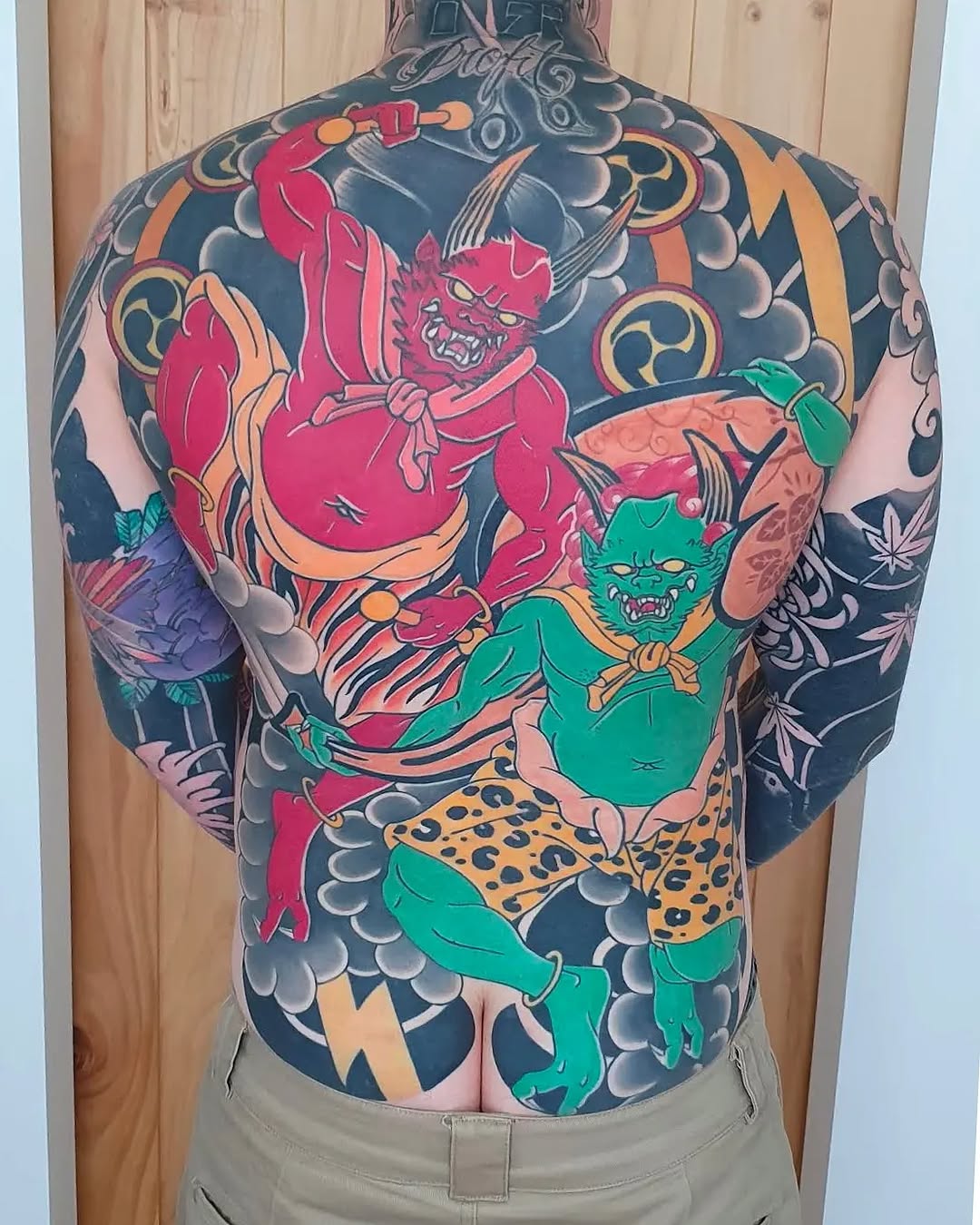 Back tattoo featuring two red and green demon figures with ornamental clouds and gold geometric patterns on dark blue background.
