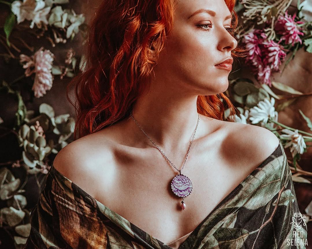 Woman with red curled hair wearing a purple flower pendant necklace and olive draped garment, posed against a floral background.