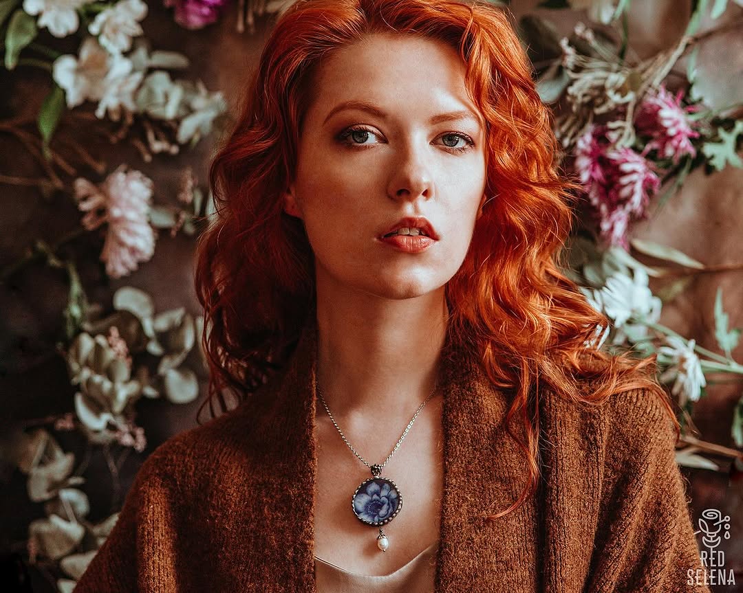 Woman with long wavy red hair wearing a brown cardigan and blue floral pendant necklace, posed against a brown background with white and pink flowers.