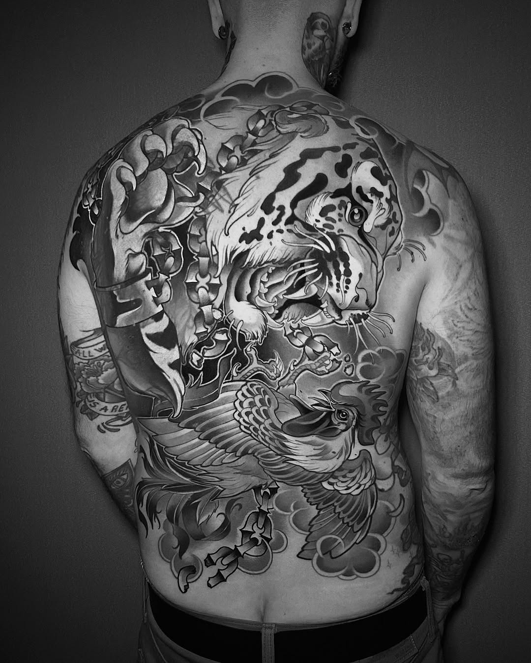 Large-scale back tattoo featuring an aggressive tiger head with ornamental clouds and floral elements in black ink on pale skin.