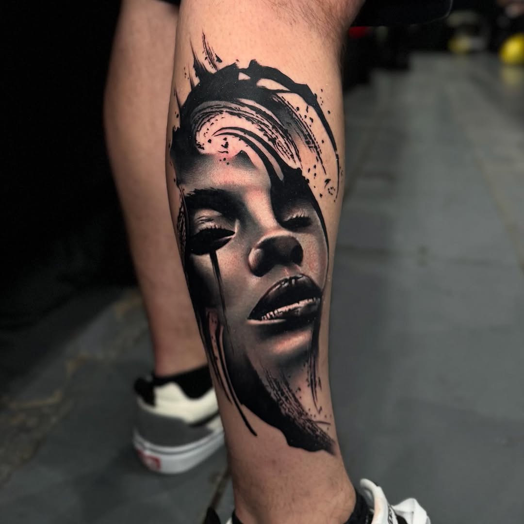 Portrait tattoo of a woman's face with closed eyes and dramatic shading on the calf, featuring black ink with splatter elements above the head.