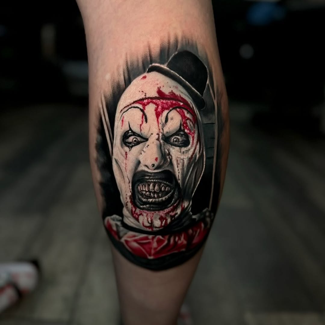 Horrifying clown face tattoo on leg featuring white makeup, red markings, black eyes and teeth, wearing a dark hood with red collar.