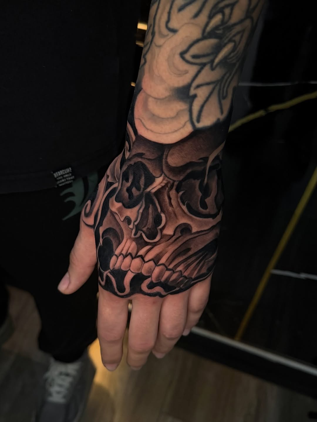Detailed skull tattoo covering the back of a hand and extending up the forearm, rendered in black and brown ink with shading and ornamental details.