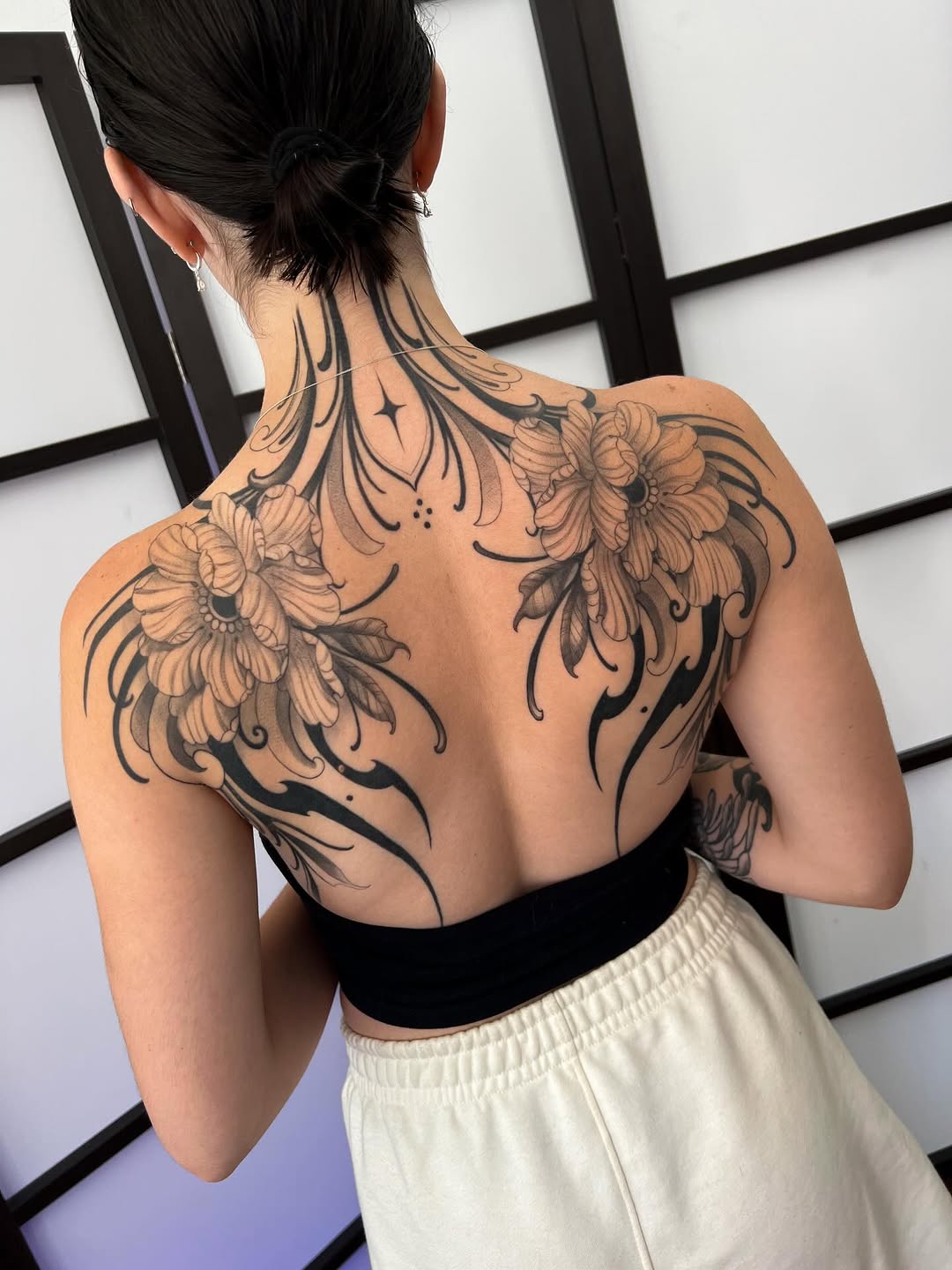 Back tattoo featuring two large flowers with flowing, curved line work extending up the neck and shoulders in black and gray ink.