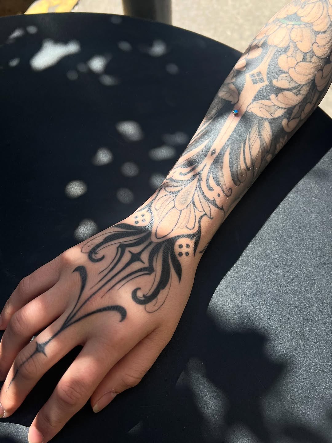 Detailed floral and ornamental arm tattoo in black ink extending from hand to upper arm, featuring layered botanical designs with scrollwork elements.