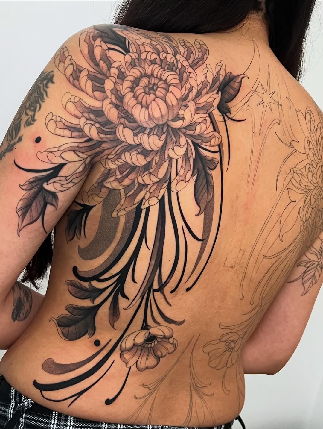 Back tattoo featuring a large chrysanthemum flower with long flowing petals in black and brown ink, with smaller floral sketches on the right side.