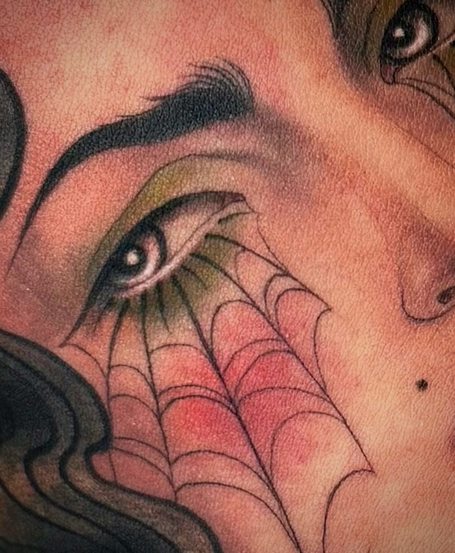 Winged eye with web design tattooed on the neck and chest area in black, green, and red ink tones.