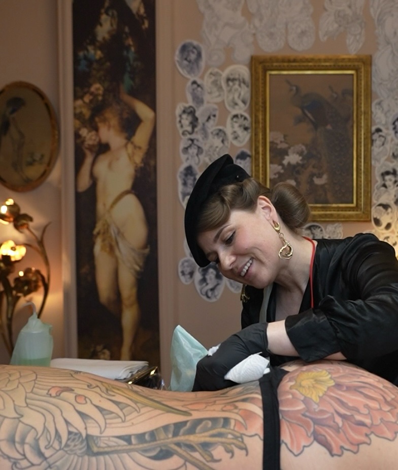 Tattoo artist applies a large colorful phoenix design on a client's back in an ornate studio setting with vintage artwork and decor.