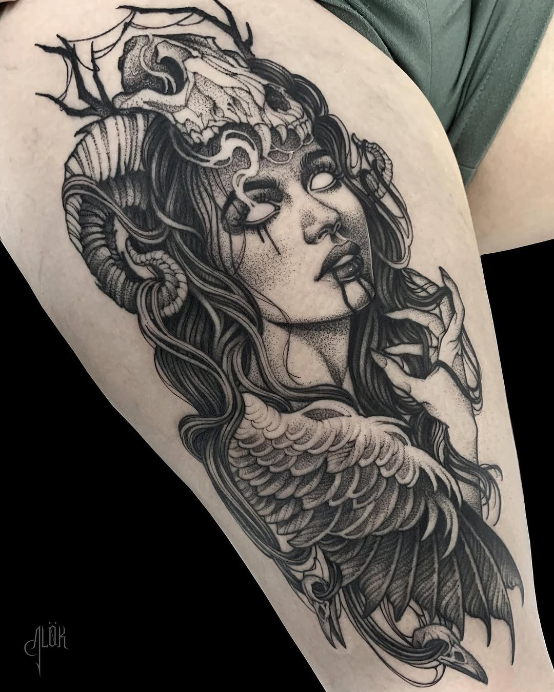 Detailed black ink tattoo on thigh featuring a winged female figure with flowing hair, ornate headpiece, and decorative feather wings.