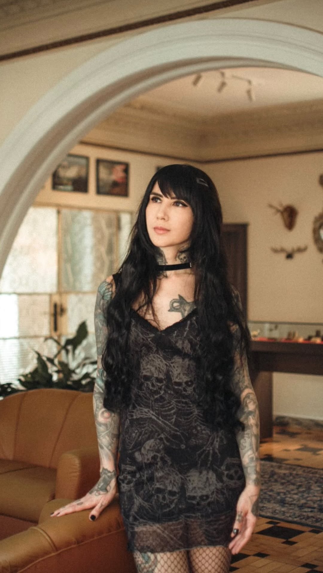 Woman with long black hair displaying extensive tattoos on both arms and torso, wearing a black lace dress in an interior space.