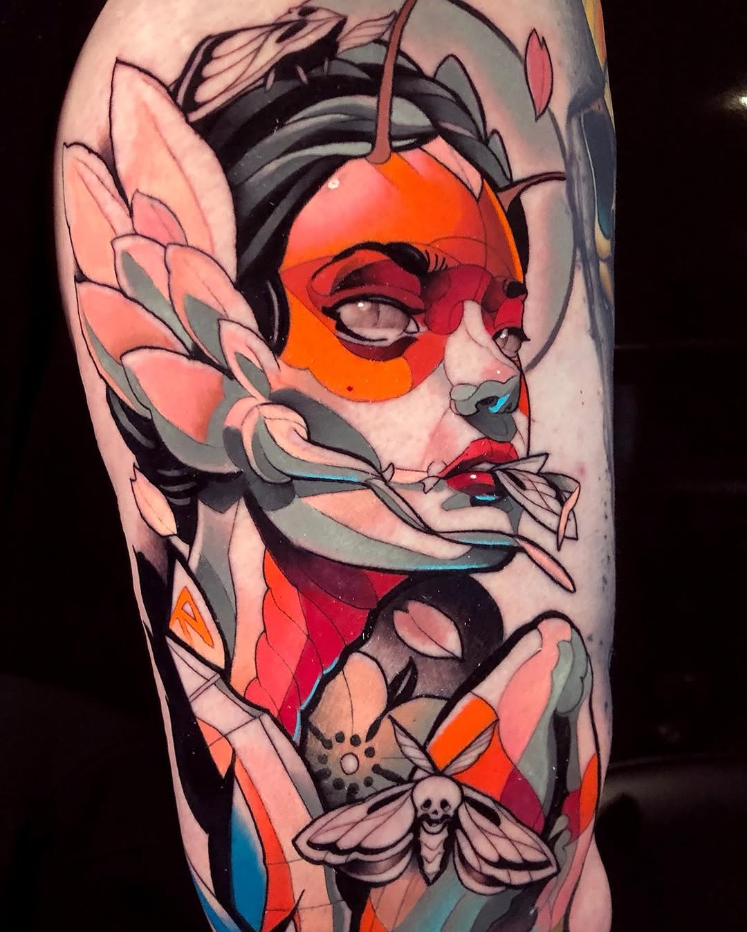 Colorful portrait tattoo on upper arm featuring a face with geometric shapes, insects, and flowers in red, orange, pink, and blue tones.
