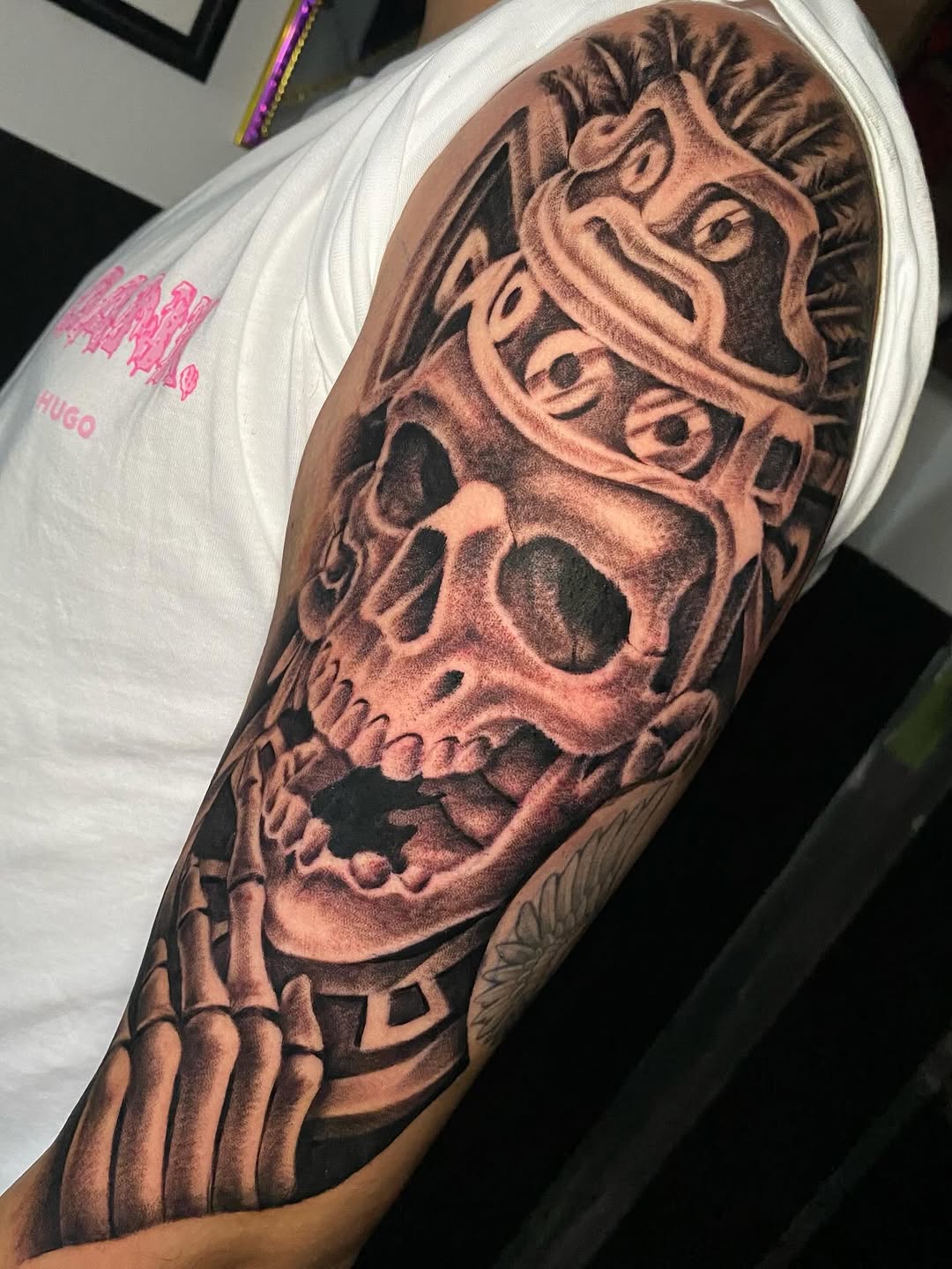 Detailed skull tattoo with skeletal face, ornamental headdress, and bone hand on upper arm in brown and black ink.