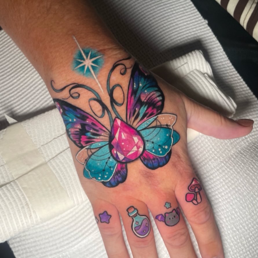 Colorful butterfly tattoo with magenta gemstone center and turquoise wings on hand, with small potion bottle and star designs on fingers.