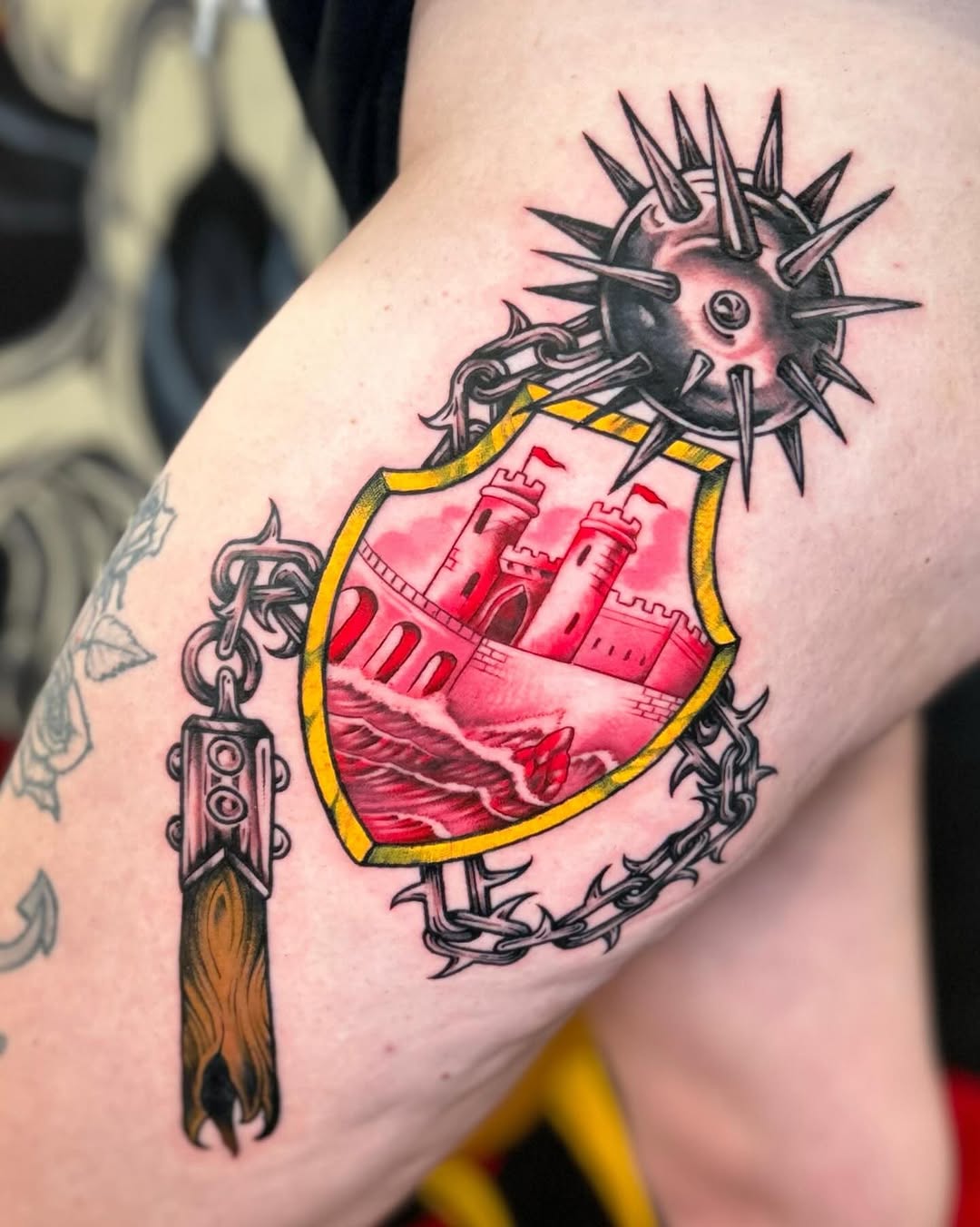 Colorful shoulder tattoo featuring a spiked mace, shield with red castle, yellow border, ornate chains, and flaming handle in red, black, and gold.