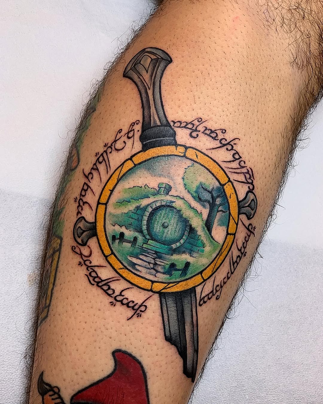 Colorful tattoo on upper arm featuring a sword with a circular gold-rimmed portal displaying a green landscape and eye, surrounded by flowing script text.