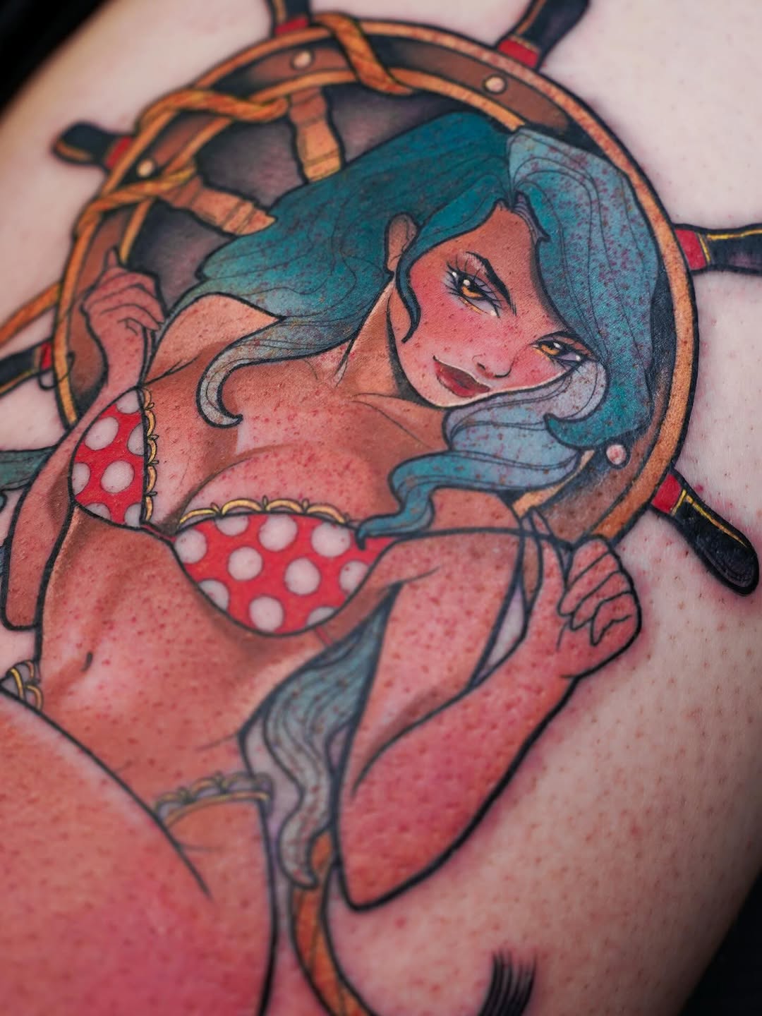 Colorful tattoo of a smiling woman with teal hair wearing a red polka dot bikini top and steering wheel, on upper arm.