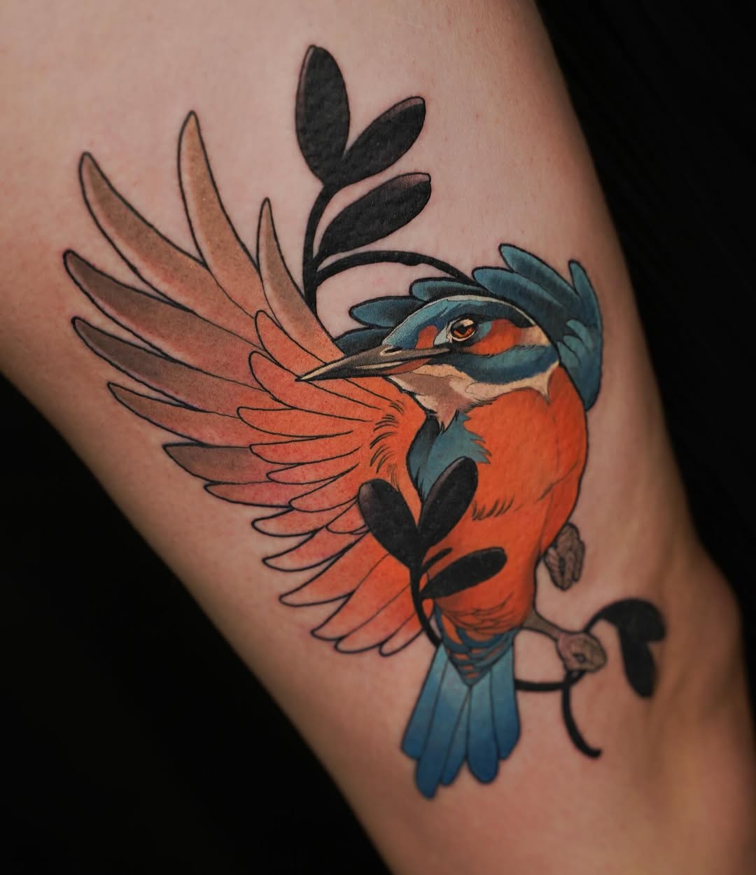 Colorful bird tattoo on thigh featuring orange breast, blue head and wings, black markings, and decorative foliage elements in vibrant hues.