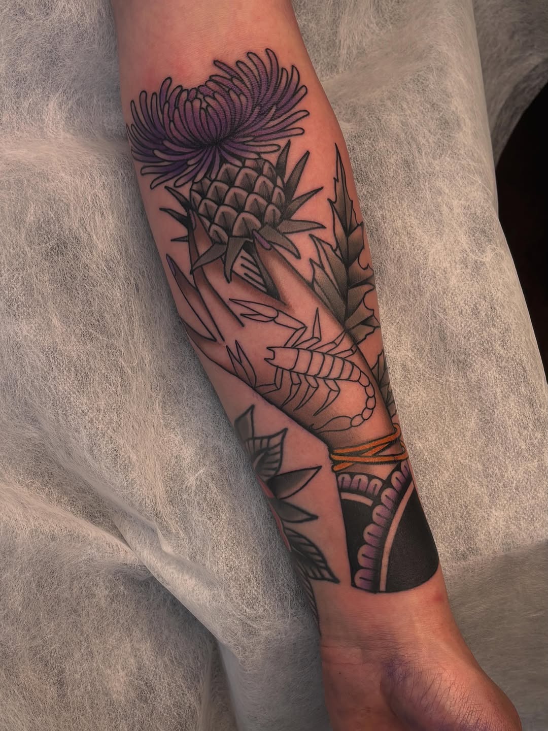 Thistle flower and leaves tattoo on forearm with purple and orange accents, featuring detailed linework and shading.